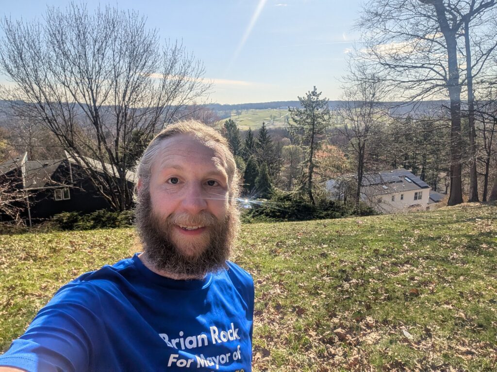 Brian standing on top of the hill, wearing a shirt that says "Brian Rock for Mayor," with the golf course in the background.